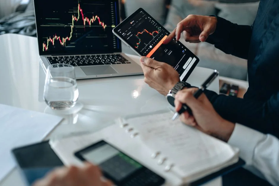 Des mains tenant un smartphone à côté d’un ordinateur portable affichant des graphiques boursiers, illustrant le trading numérique.